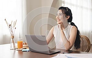 Relax young asian businesswoman looking out the window and working on laptop computer while sitting at the kitchen room background