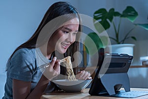 Relaxation, alone smile asian young woman eating instant ramen, noodles while using, watching video, movie, media on pc, tablet