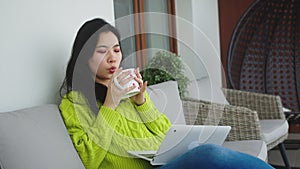 Relaxation and wellbeing concept. Young asian woman drinking cup of tea while resting on the balcony sofa with laptop