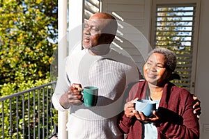 Relaxed african american senior couple drinking coffee standing on balcony in sun