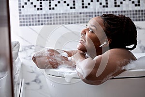 Relaxed african woman taking bath with foam with pleasure, enjoying spa, hygiene and listen to music