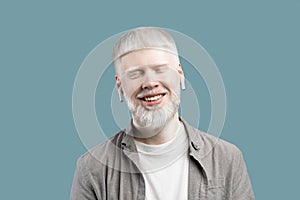 Relaxed albino guy listening music in bluetooth earpods, standing with closed eyes over turquoise background