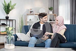 Relaxed Arabian Muslim couple in casual wear, using a laptop pc together for watching online content or shopping