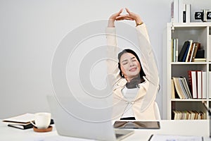Relaxed Asian businesswoman sits at her desk and stretches her arms after finishing work