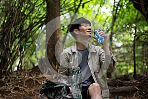 A relaxed Asian male hiker is sitting, drinking water, and taking a break during his hiking trip