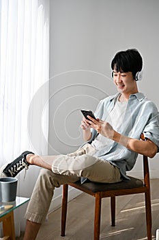 Relaxed Asian man istening to music through his headphones, using smartphone, sitting on a chair
