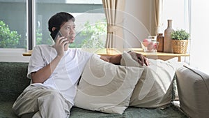 Relaxed asian man sitting on comfortable sofa at home and having conversation with mobile phone.