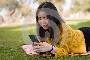 Relaxed Asian woman using mobile phone in city park - lifestyle portrait of young happy and pretty Chinese girl lying on green