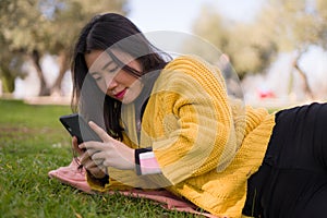 Relaxed Asian woman using mobile phone in city park - lifestyle portrait of young happy and pretty Japanese girl lying on green