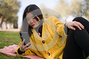 Relaxed Asian woman using mobile phone in city park - lifestyle portrait of young happy and pretty Korean girl lying on green