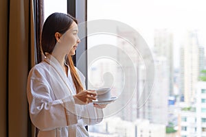 Relaxed Asian woman wearing bathrobe drinking coffee in hotel room