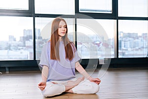 Relaxed attractive young woman meditating sitting on floor in lotus pose on background of window in light office room.