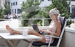 Relaxed barefoot senior woman sitting and typing on a laptop computer outside on a balcony