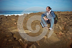 Relaxed bearded male traveler, adventurer, backpacked tourist sitting on a rocky cliff, looking into the distance