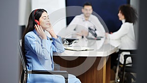 Relaxed beautiful Asian woman in headphones dancing sitting at conference table with blurred colleagues talking at