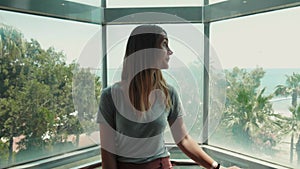 Relaxed beautiful woman smiling at the opening view of the sea in transparent elevator going up on tropical vacation.