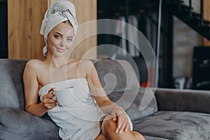 Relaxed beautiful young woman has healthy skin wears soft towel on head and around naked body holds mug of coffee poses at sofa