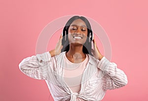 Relaxed black woman in headphones enjoying good song and smiling with closed eyes on pink studio background