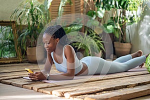 Relaxed black woman lying on fitness mat holding smartphone take break after home sported training.