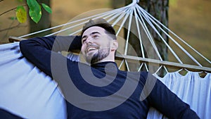 Relaxed brunette young man with blue eyes looking up lying on hammock smiling. Portrait of carefree satisfied Caucasian