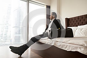 Relaxed businessman sitting on bed besides luggage bag.