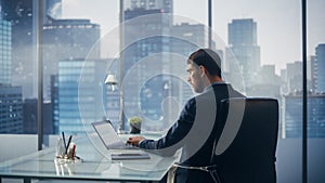 Relaxed Businessman in a Suit Sitting at a Desk in Modern Office, Using Laptop Computer, Next to