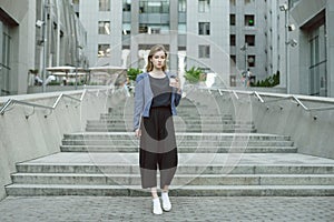 Relaxed businesswoman standing near office building holding laptop, smartphone and coffee. Horizontal shot of female office worker