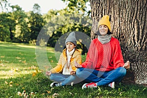 Relaxed carefree woman and little daughter sit in pose of lotus near tree in park, close eyes, try to concentrate, practice yoga t