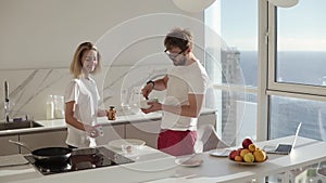 Relaxed caucasian man preparing breakfast for two in kitchen at home and mixing eggs in bowl, blonde girl preparing