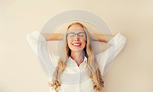 Relaxed caucasian middle-aged woman manager resting with closed eyes and folded hands behind the head in eyeglasses on white wall