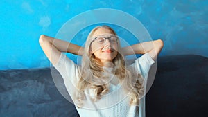 Relaxed caucasian middle-aged woman resting with closed eyes and folded hands behind the head in eyeglasses while sitting on couch