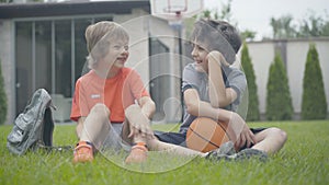 Relaxed Caucasian schoolboys sitting on green grass at home and talking. Portrait of joyful boys with basketball ball
