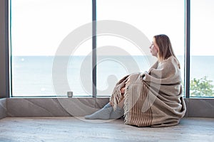 Relaxed Caucasian woman sitting on a warm floor in a socks wrapped in a woolen blanket near the large window in light. autumn mood