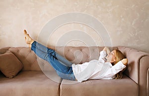 Relaxed caucasian young woman resting with closed eyes and folded hands behind the head while lying on couch in the living room at