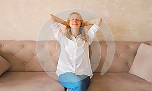 Relaxed caucasian young woman resting with closed eyes and folded hands behind the head while sitting on couch in the living room