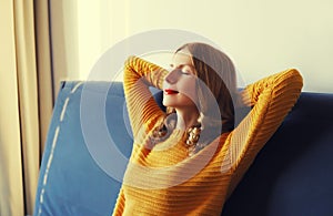 Relaxed caucasian young woman resting with closed eyes and folded hands behind the head sitting on the couch in room at home