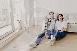 Relaxed couple sit on floor near couch, embrace and smile, dressed in casual clothes and white socks, enjoy domestic atmosphere,