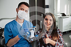Relaxed at the dentist. Happy female patient sitting in a dental chair smiling to the camera with her dentist holding false tooth