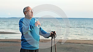 Relaxed elderly man with Scandinavian walking stick drinking water bottle at sea sand beach sunset