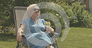 Relaxed elegant senior woman sitting in chair outdoors and drinking water. Portrait of carefree Caucasian lady in