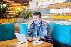 Relaxed entrepreneur wearing protective face mask sitting at table in coffee shop looking at digital tablet while talking on