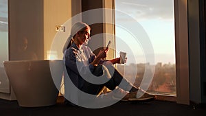 Relaxed female sitting in corner with cell phone and plastic coffee cup