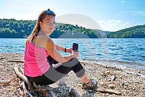 Relaxed female tourist with a thermal insulated coffee mug in hands, sitting down on a log at a mountain lake shore.