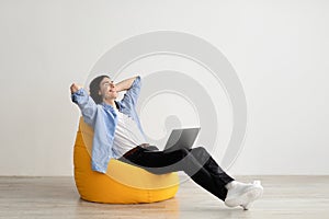 Relaxed freelancer guy leaning back in beanbag chair, resting after work on laptop, sitting with hands behind head