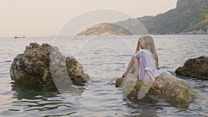 Relaxed girl in white tunic enjoying sea landscape on stone in water. Happy young girl sitting on rock in sea water on