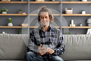 Relaxed Guy Wearing Headphones Using Mobile Phone Sitting At Home
