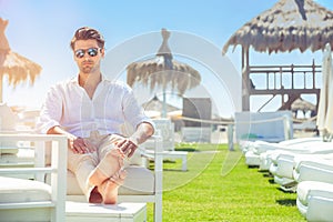 Uvolněná pěkný muž na bílém židle během na pláž. bílý košile a sluneční brýle. nohy protáhl ven 
