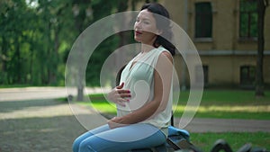 Relaxed happy Caucasian pregnant woman sitting on bench in city stroking belly in slow motion thinking. Side view