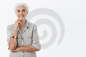 Relaxed and happy charming old lady with grey hair in casual shirt and bracelet leaning head on fist above chin smiling