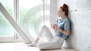 Relaxed happy redhead young woman drinking coffee while gazing out of window.